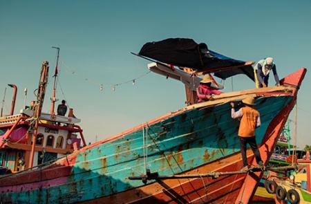 A fishing vessel undergoing maintenance in Juwana, Pati Regency, Central Java, Indonesia.  Two people are on board, and one person stands on a platform below the ship. Photo by Alfi Hilman for Unsplash.