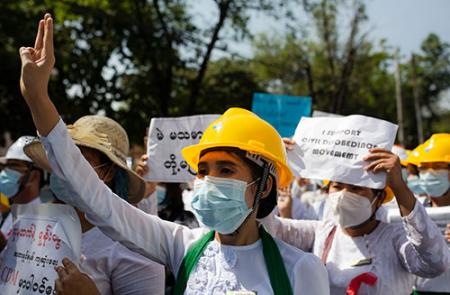 A women wearing a hard hat and surgical mask stands among a crowd of workers with her hand in the air. Many of the workers are carrying signs.