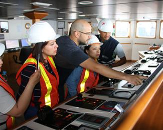 Five workers in safety helmets and reflective vests are operating and monitoring equipment in a ship's control room.