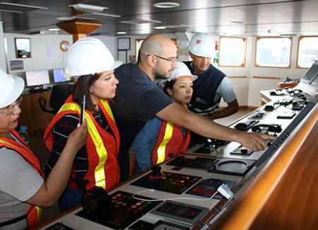 Five workers in safety helmets and reflective vests are operating and monitoring equipment in a ship's control room.